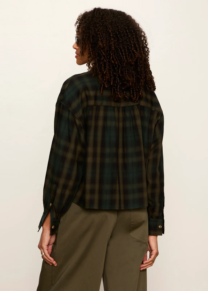 A woman shown from the back wearing a dark green and black plaid long-sleeve shirt with olive green pants, photographed against a plain off-white studio background.