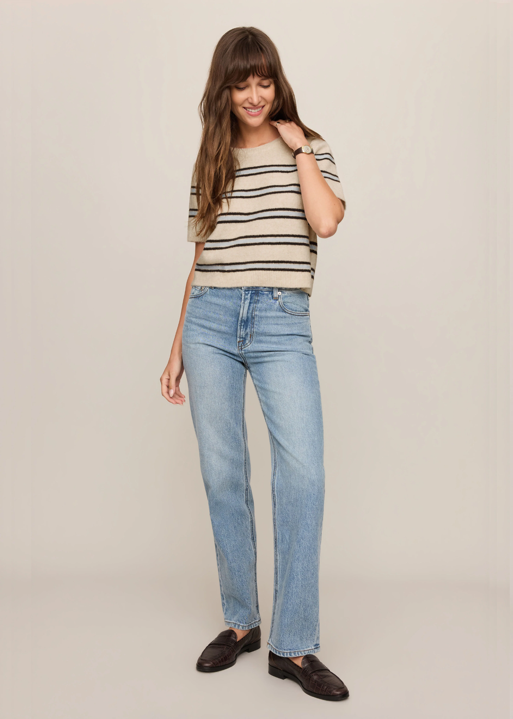 A woman modeling light-wash blue straight-leg jeans, a beige and navy striped knit top, and dark brown leather loafers, standing against a plain beige studio background.