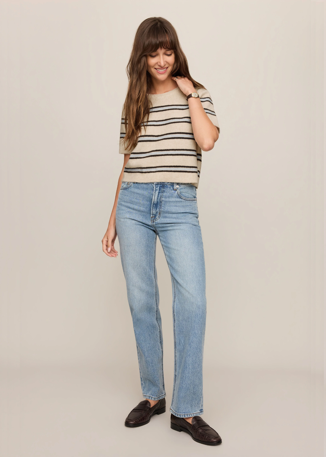 A woman modeling light-wash blue straight-leg jeans, a beige and navy striped knit top, and dark brown leather loafers, standing against a plain beige studio background.