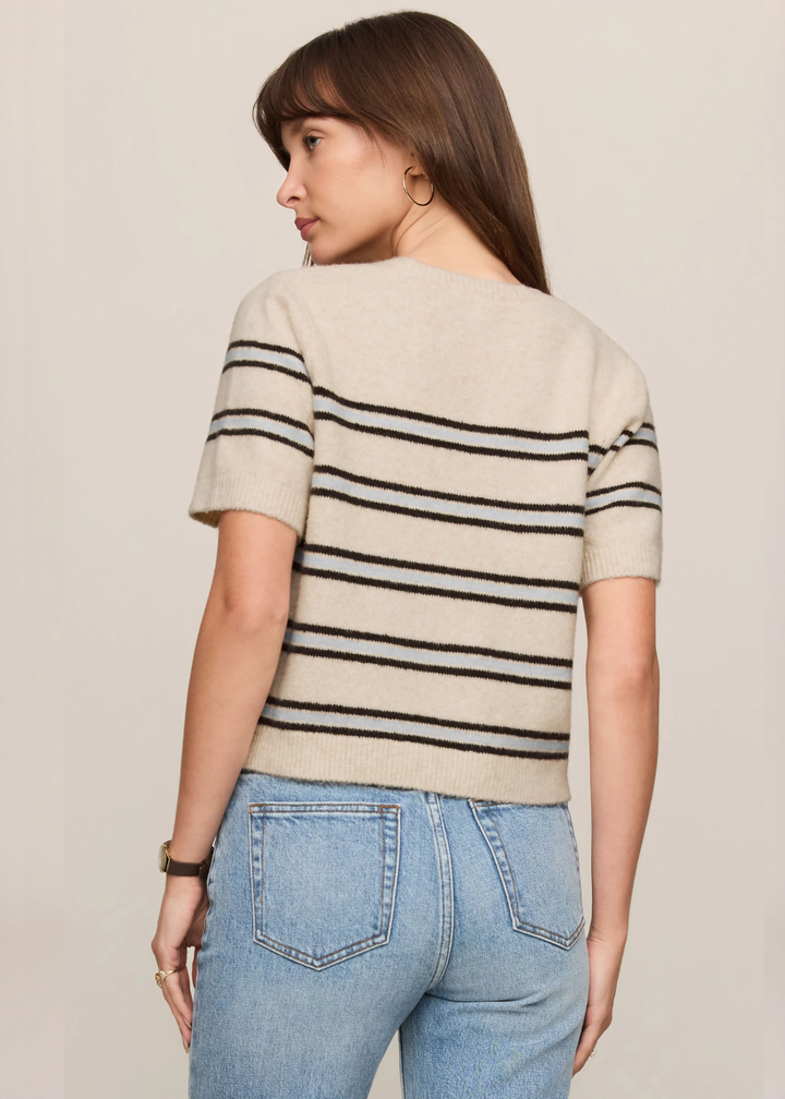 A woman shown from the back wearing a short-sleeve knit sweater with horizontal black stripes on a cream base, paired with light blue jeans, against a plain beige studio background.