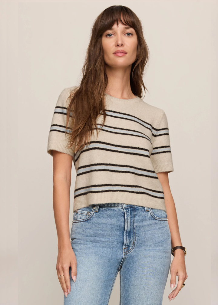 A woman modeling a short-sleeve knit top with horizontal black stripes on a beige background, paired with light blue jeans, against a plain beige studio backdrop.