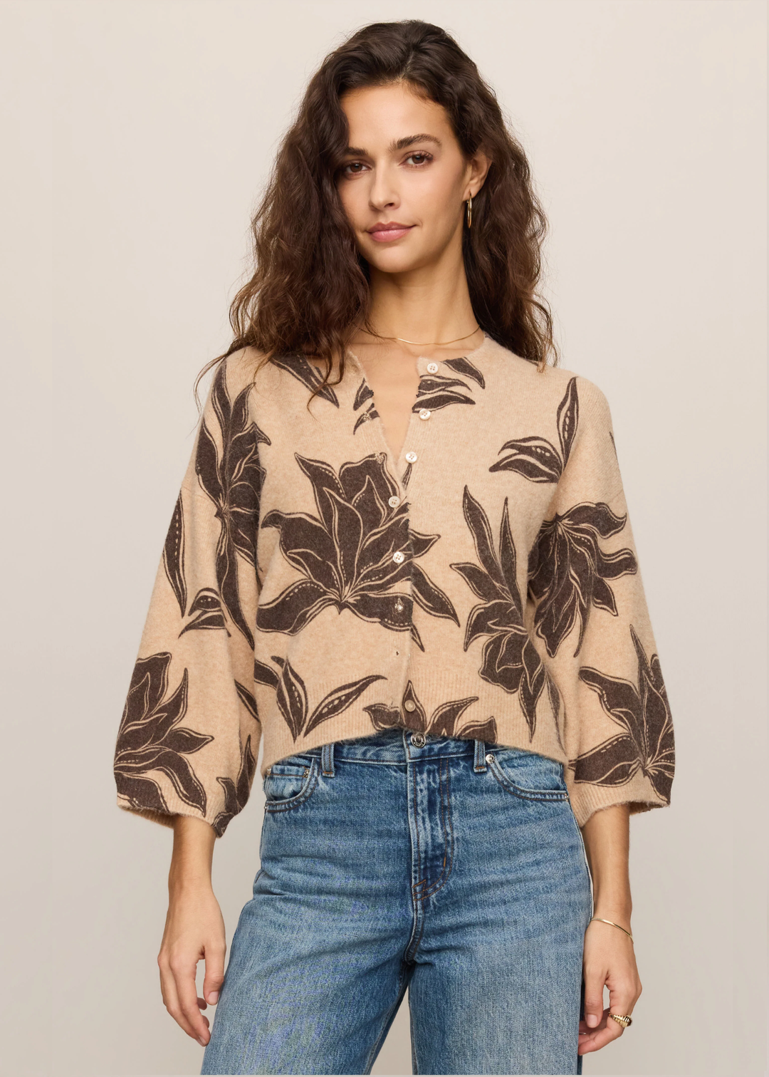 A woman modeling a beige button-up blouse with a dark brown tropical floral pattern, paired with blue jeans, against a solid light beige background.