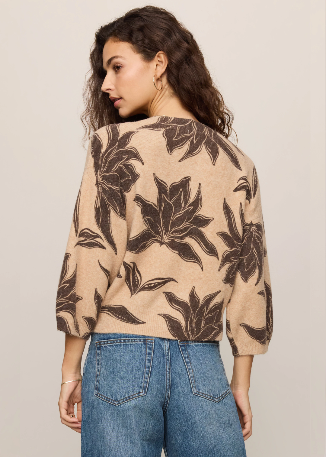 A woman shown from the back wearing a beige cropped top with a dark brown floral pattern, paired with blue jeans, against a plain light beige background.
