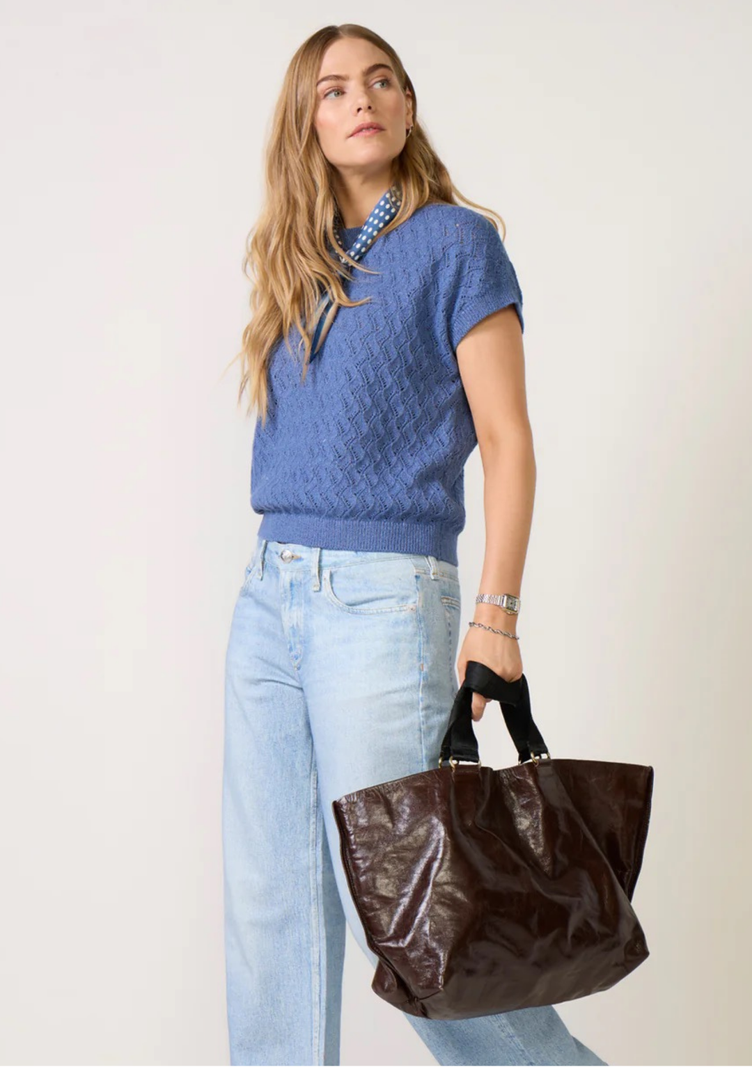 Woman wearing a blue sweater and light blue jeans, holding a brown bag against a white background