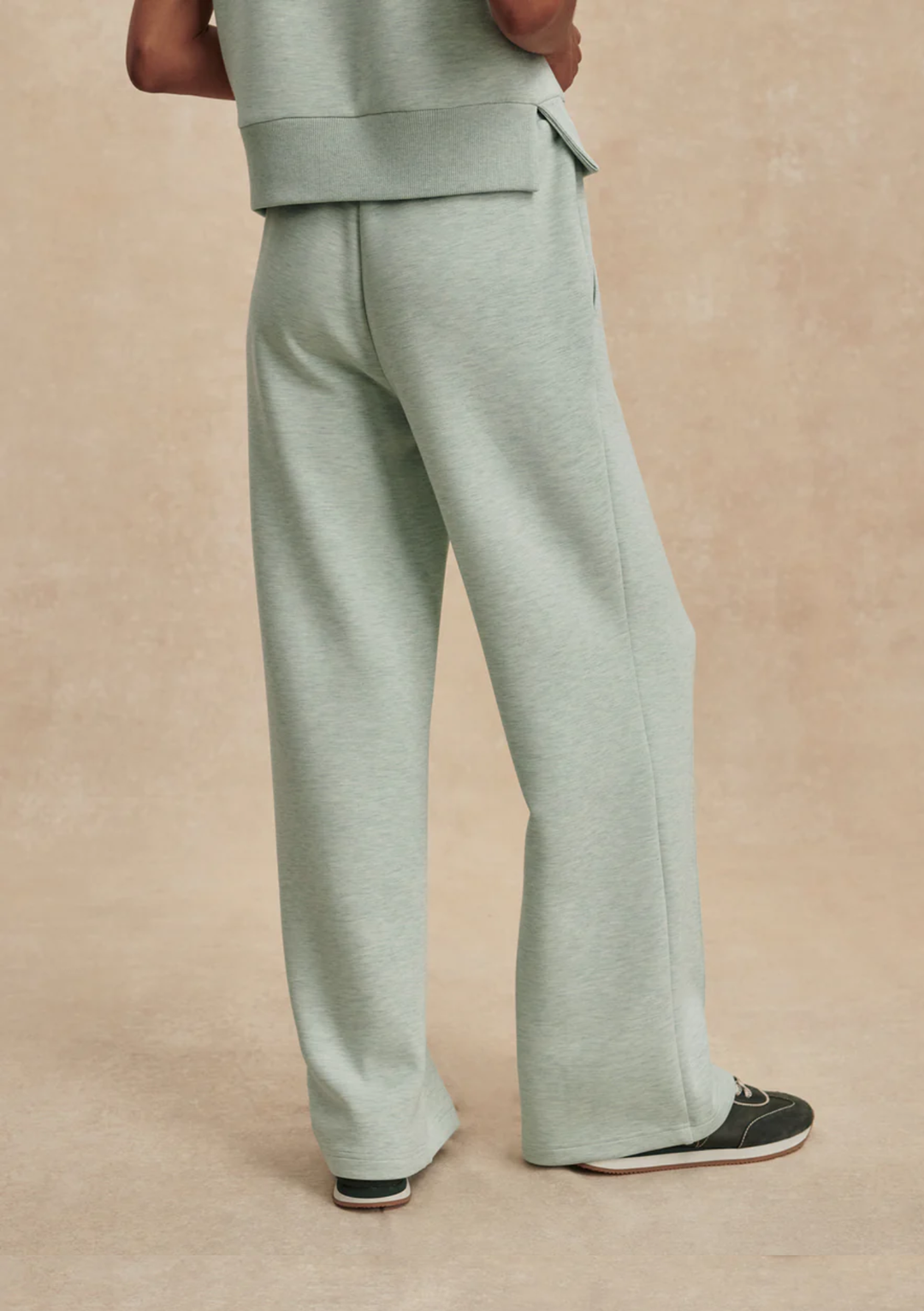 A person shown from the back wearing light sage green wide-leg sweatpants and a matching top, standing against a warm beige studio backdrop.