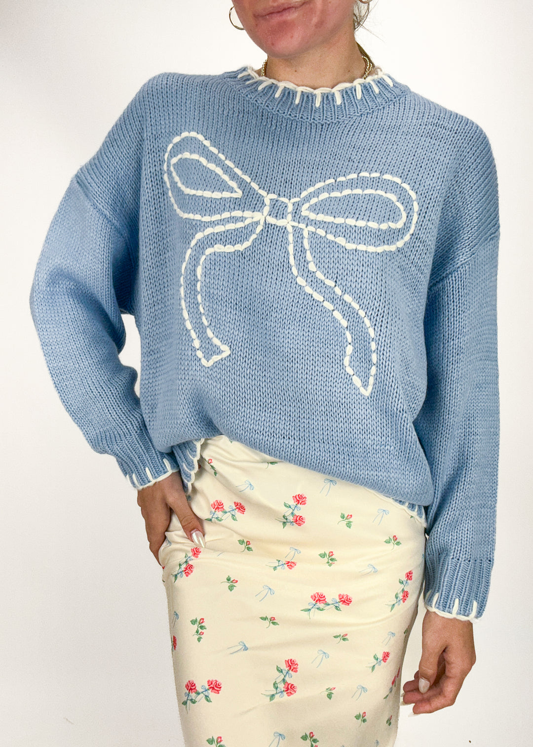 woman wearing a light blue knit sweater with white embroidered bow on chest and whipstitch details at collar, sleeves, and hem