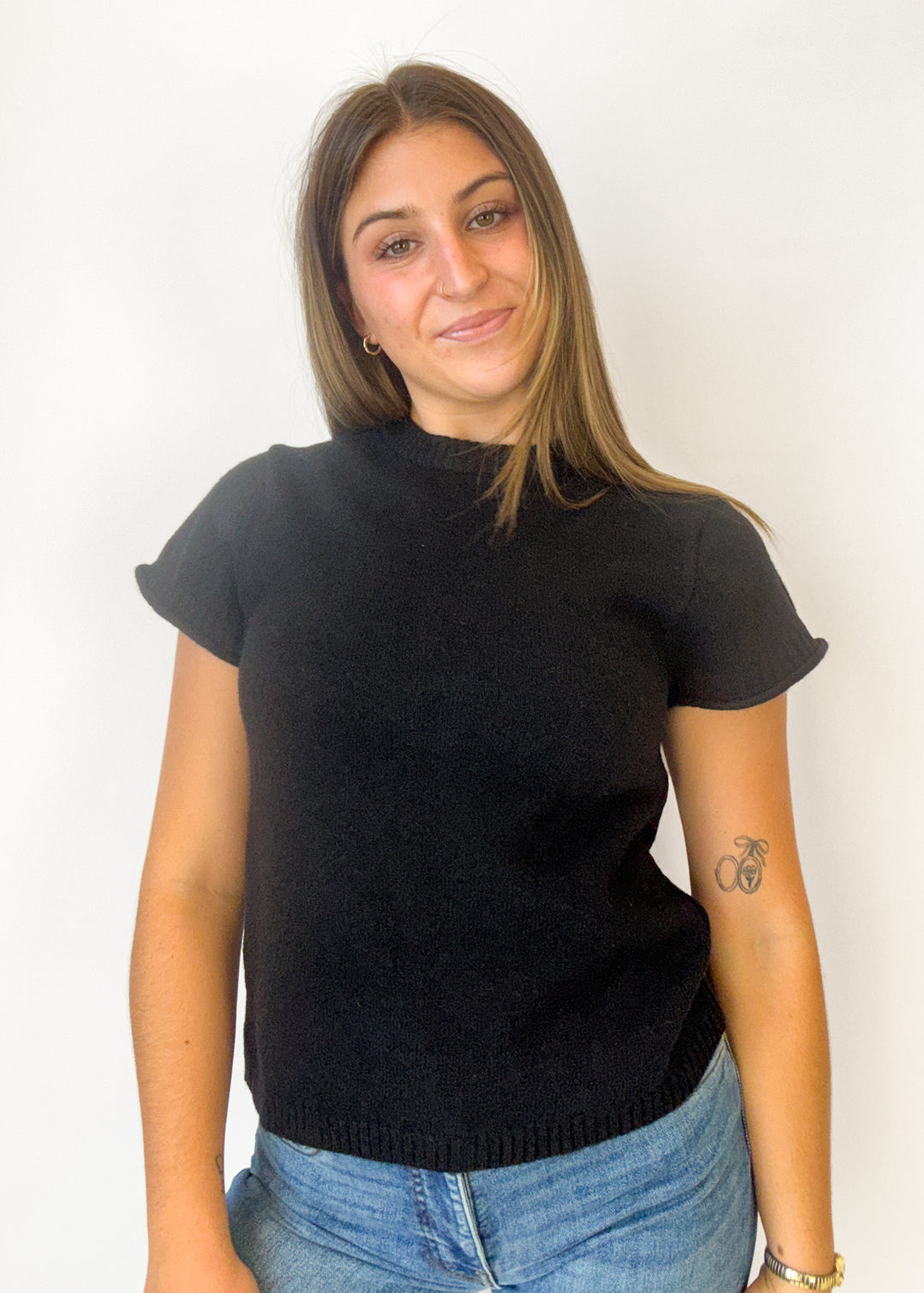 A woman wearing a black short-sleeve knit sweater and blue jeans, standing against a plain white wall.