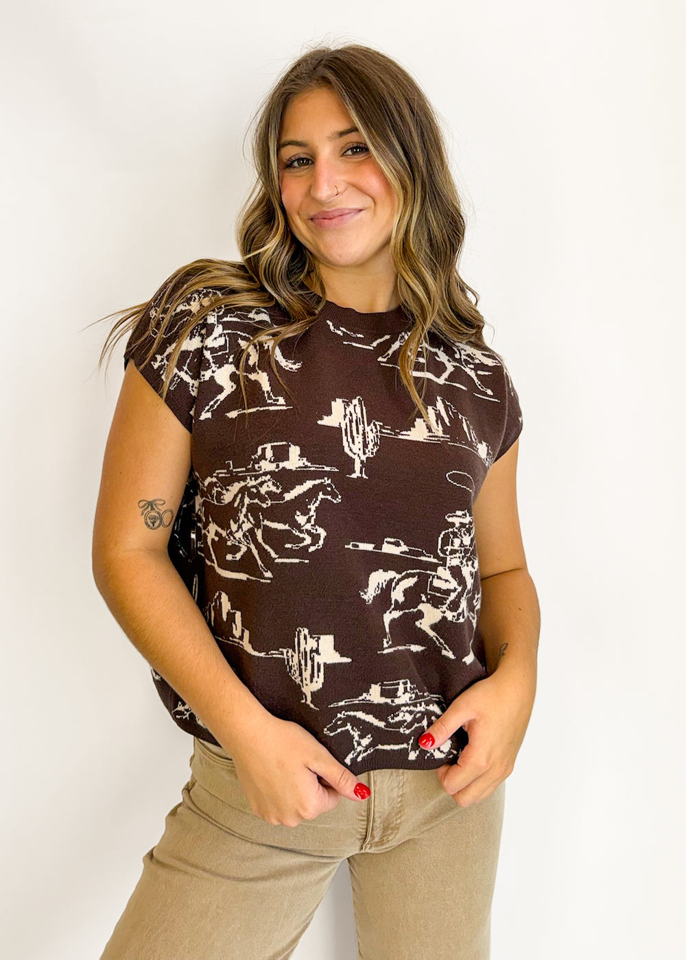 A woman wearing a brown short-sleeve t-shirt with a white western cowboy print, paired with beige pants, standing against a plain white background.