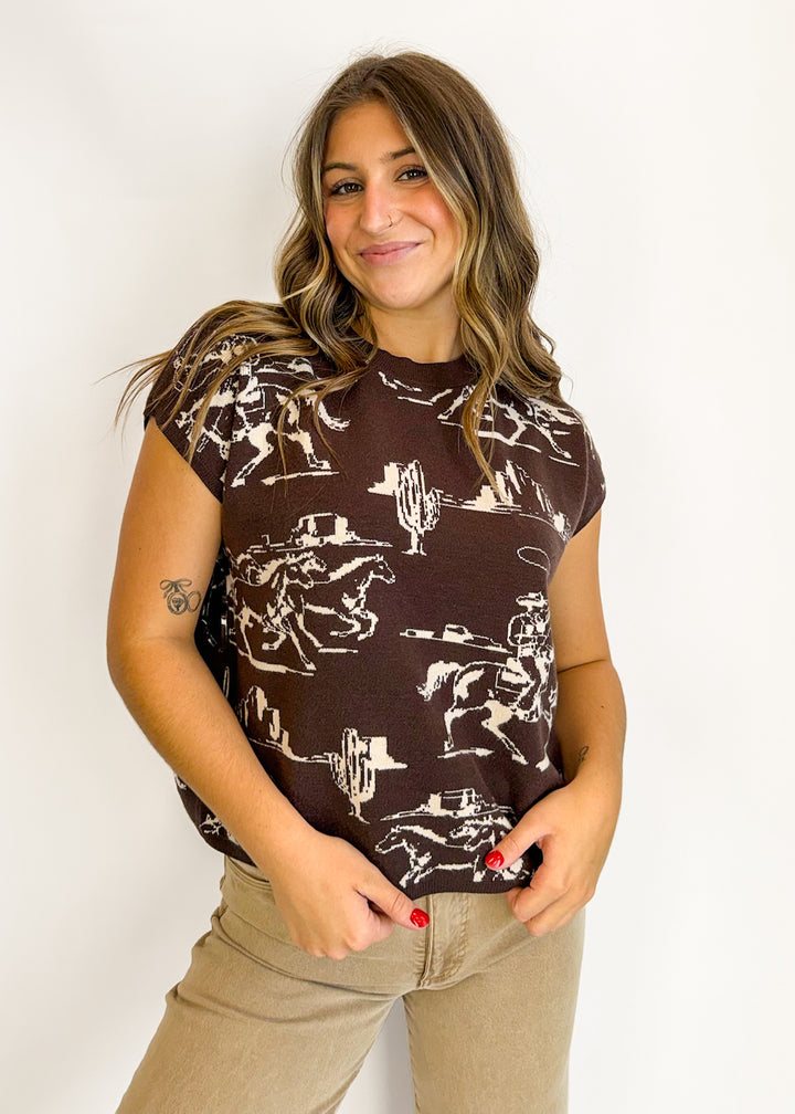A woman wearing a brown short-sleeve t-shirt with a white western cowboy print, paired with beige pants, standing against a plain white background.