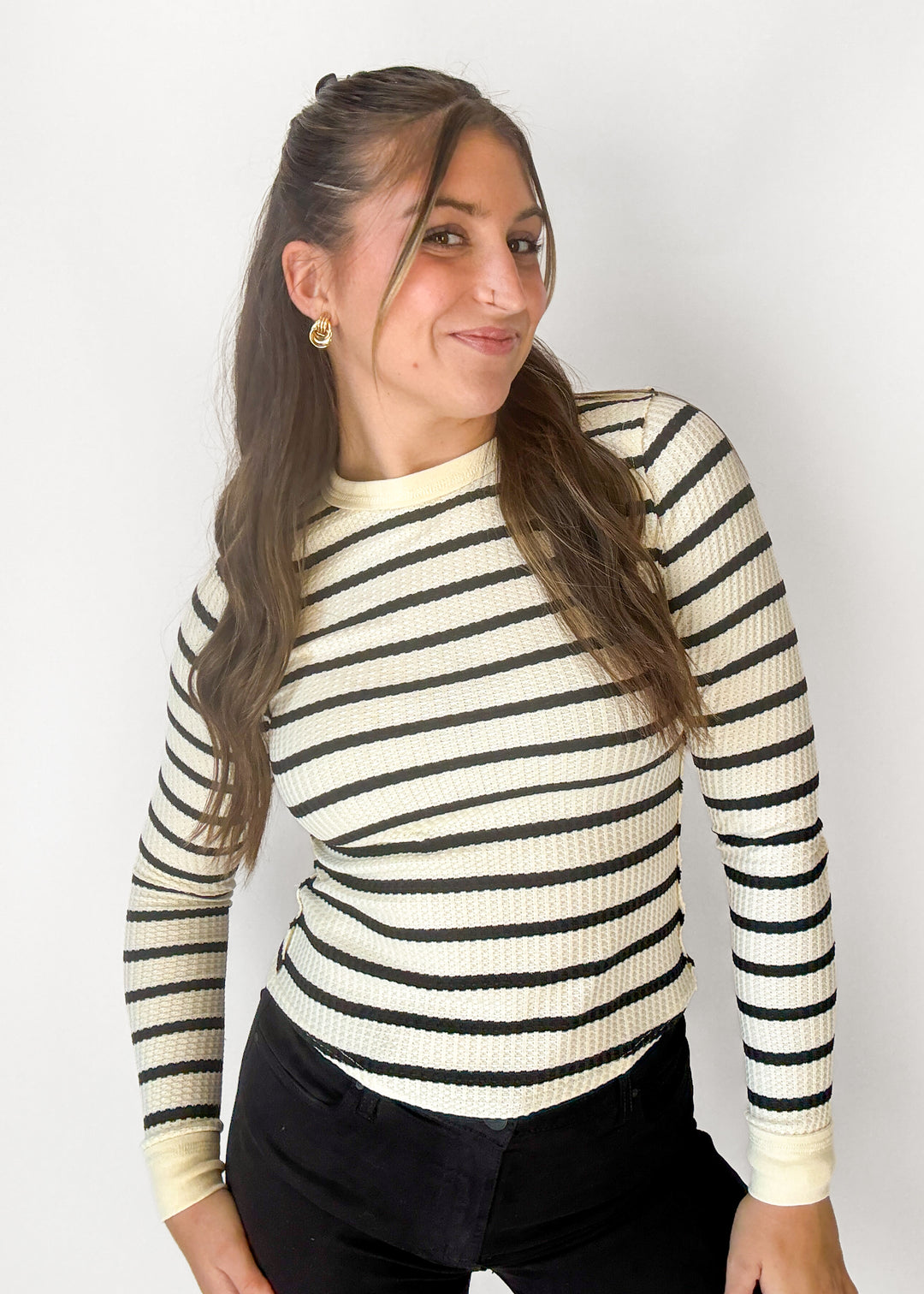 A woman wearing a cream and black horizontally striped long-sleeve knit top, paired with black pants, standing against a plain light gray background.
