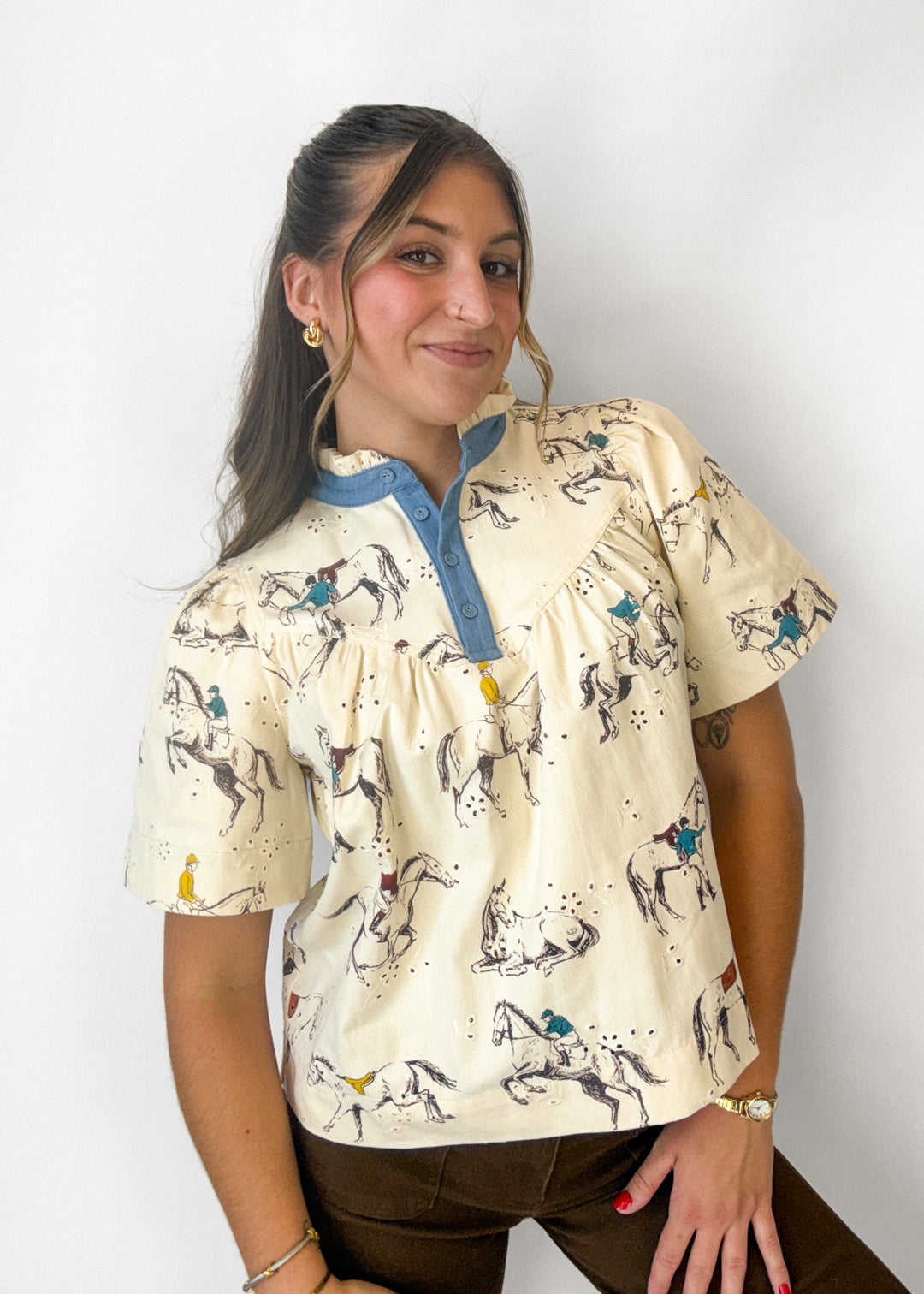 A woman wearing a cream-colored short-sleeve blouse with a blue collar and placket, decorated with a western horse print, paired with brown pants, set against a plain white background.