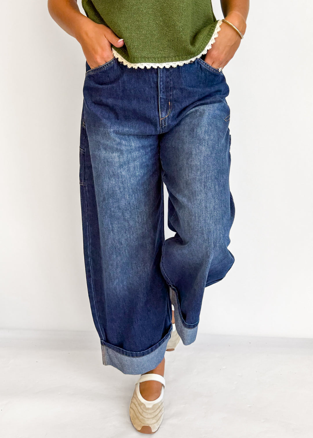 A person wearing loose, wide-leg blue denim jeans with rolled cuffs, paired with a green top and beige wedge sandals, standing against a plain white background.