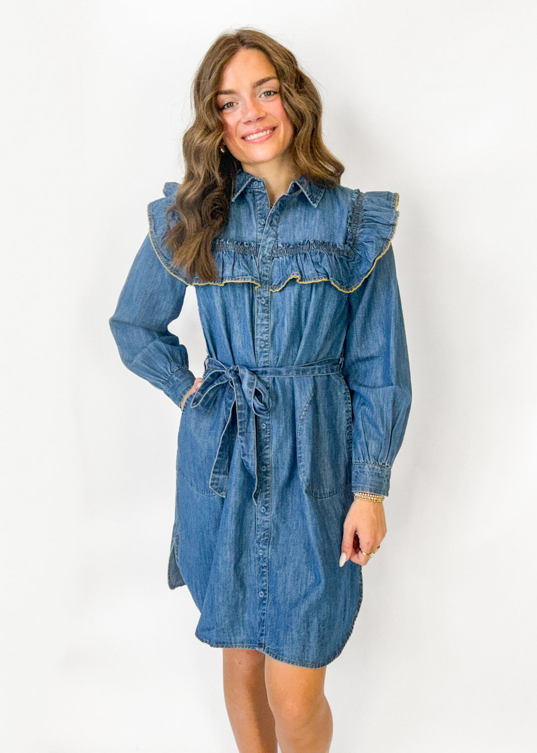 A woman modeling a blue denim shirt dress with a large ruffled collar, button front, long sleeves, and a self-tie waist belt, photographed against a plain white studio background.