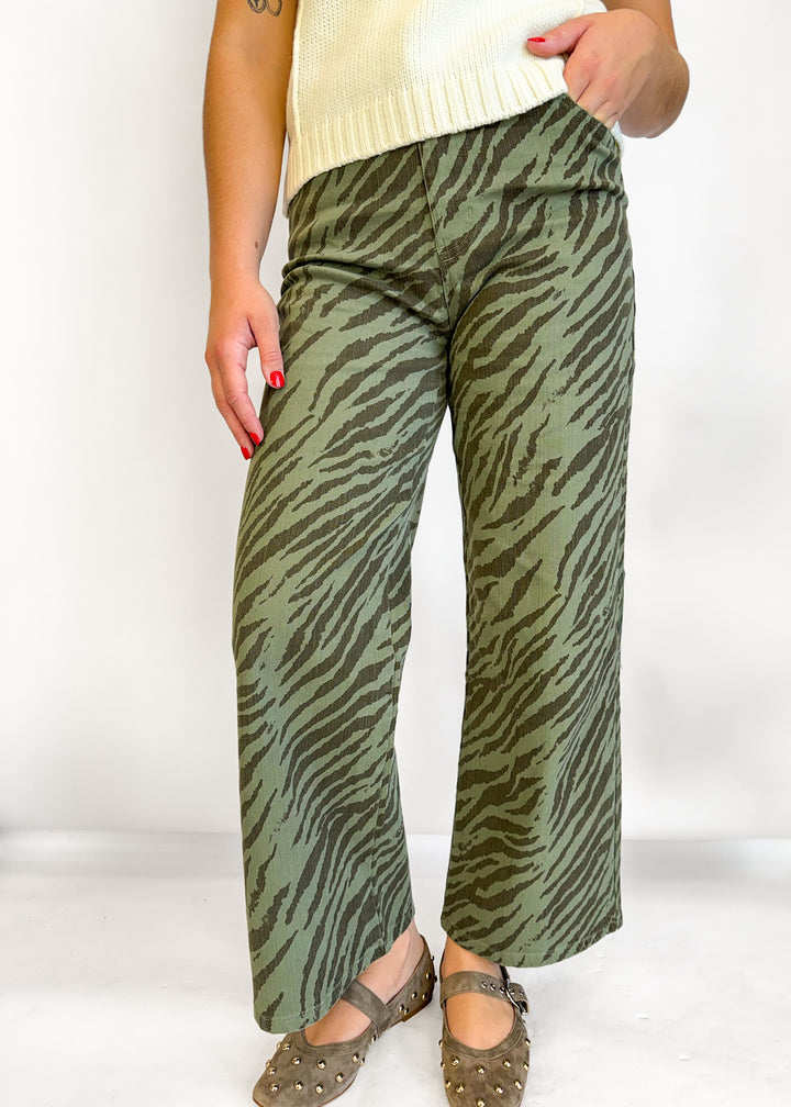 A person wearing olive green wide-leg pants with a dark brown zebra stripe pattern, styled with a cream ribbed knit top and brown studded flat shoes against a white studio background.