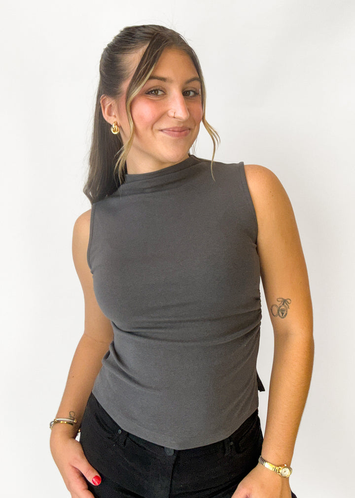 A woman wearing a dark gray sleeveless mock neck top and black pants, standing against a plain white background.
