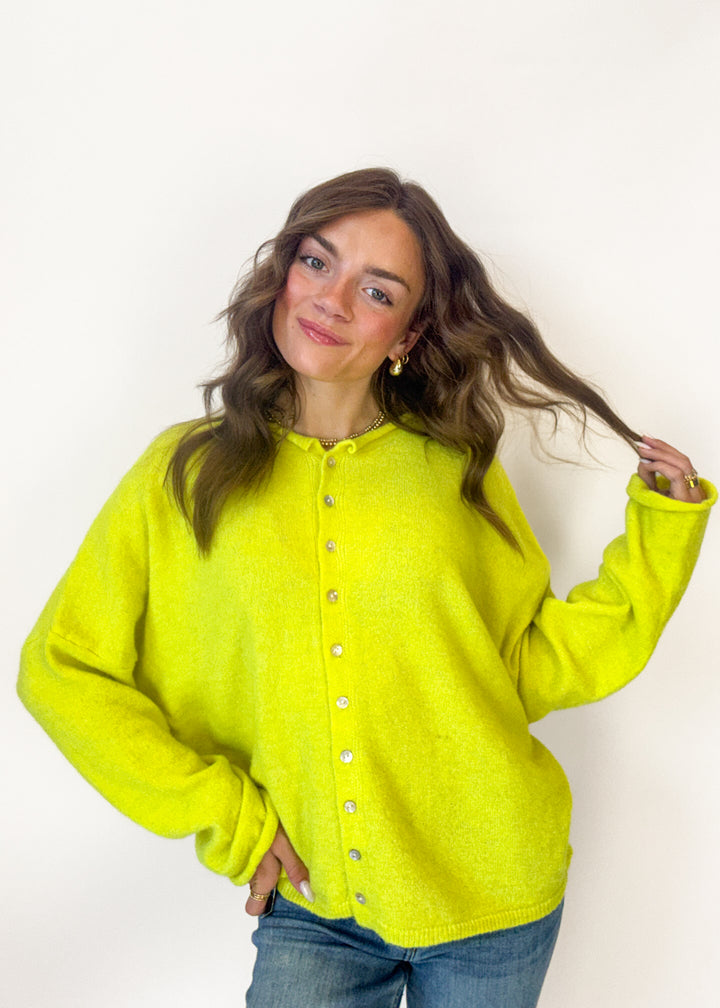 A woman wearing a bright neon yellow-green fuzzy button-up cardigan sweater and blue jeans, posing against a plain white background.