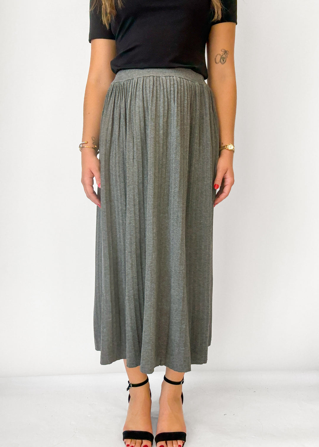 A woman wearing a high-waisted, pleated gray maxi skirt with a black short-sleeve top and black high-heeled sandals, standing against a plain white studio background.