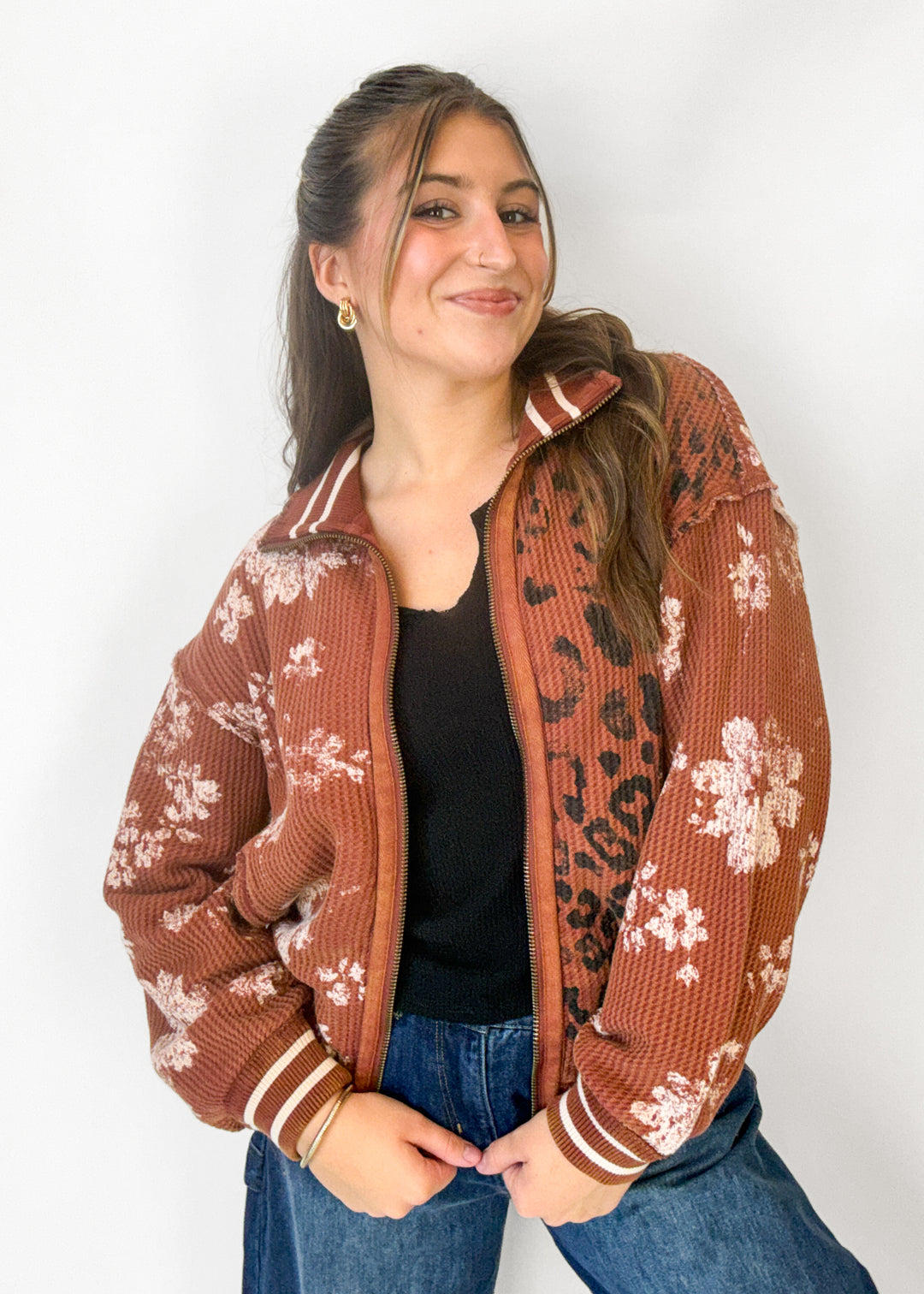 A woman wearing a rust-brown waffle-knit zip-up jacket with white floral and black leopard print patterns, styled with a black top and blue jeans against a plain white background.