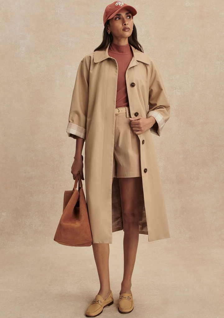 Woman wearing a beige trench coat and shorts with a brown bag against a beige background