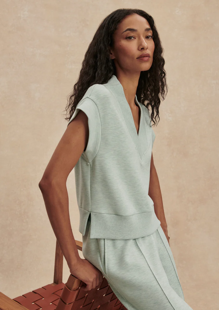 A woman wearing a light sage green sleeveless knit top and matching pants, posed next to a brown woven chair against a textured beige background.